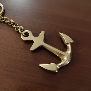 Op de afbeelding: Een goudkleurige anker sleutelhanger met een ketting. Het anker heeft een klassiek ontwerp met een ring aan de bovenkant en een gebogen bodem. De sleutelhanger is gemaakt van metaal en wordt waarschijnlijk gebruikt om sleutels te bewaren.
