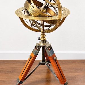 Op de afbeelding: Een messing armillarium met concentrische ringen gegraveerd met sterrenbeelden zoals Vissen en Waterman, gemonteerd op een houten driepootstatief met verstelbare metalen onderdelen en een centrale verticale steunkolom.