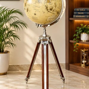 May include: A floor-standing world globe featuring a vintage-style map with beige oceans and dark landmasses. The globe is mounted in a polished silver-tone metal meridian and rests on a tripod base made of dark brown wood with silver metal accents.