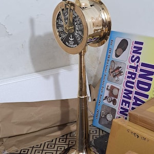 May include: A brass engine room telegraph with a black and gold dial, displaying directional instructions and the word "TELEGRAPH." It has wooden handles and is mounted on a brass stand with a dark wooden base. The dial indicates "SLOW," "HALF," and "FULL."