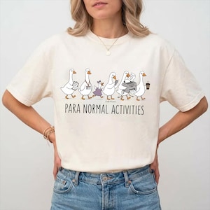 Puede incluir: Una camiseta de manga corta color crema con un gráfico de cinco gansos blancos realizando tareas de oficina. Debajo de los gansos, el texto dice PARA NORMAL ACTIVITIES en letras mayúsculas negras. Los gansos aparecen con varios objetos.