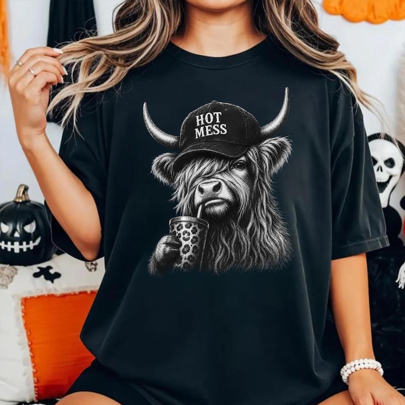 May include: A black short-sleeve t-shirt featuring a graphic of a shaggy highland cow wearing a black baseball cap with the text "HOT MESS" printed in white. The cow is depicted holding a leopard-print cup with a straw in a monochromatic style.