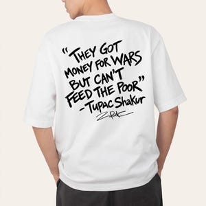 May include: A white short-sleeved t-shirt featuring black text on the back that reads: "THEY GOT MONEY FOR WARS BUT CAN'T FEED THE POOR" - Tupac Shakur, followed by a stylized signature. The lettering appears in a bold, hand-written brush style.