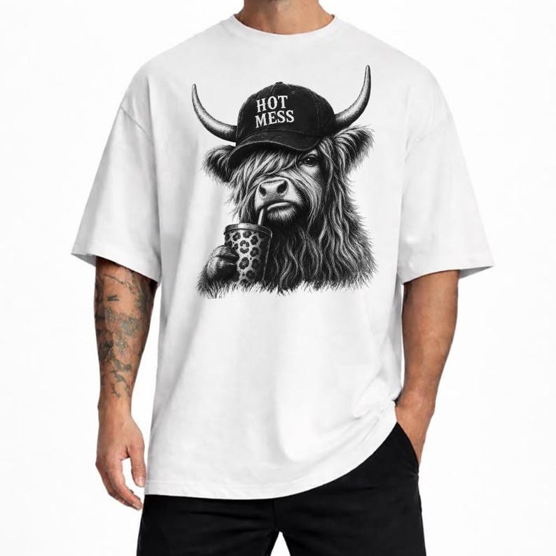 May include: A white t-shirt featuring a black and white graphic of a shaggy Highland cow wearing a baseball cap with the text HOT MESS printed on it. The cow is holding a cup with a leopard print pattern and a straw in its mouth.