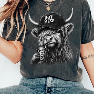 May include: A charcoal gray t-shirt featuring a graphic of a shaggy Highland cow wearing a black baseball cap with the text HOT MESS printed in white. The cow is holding a tumbler with a leopard print pattern. The shirt has a vintage wash finish.