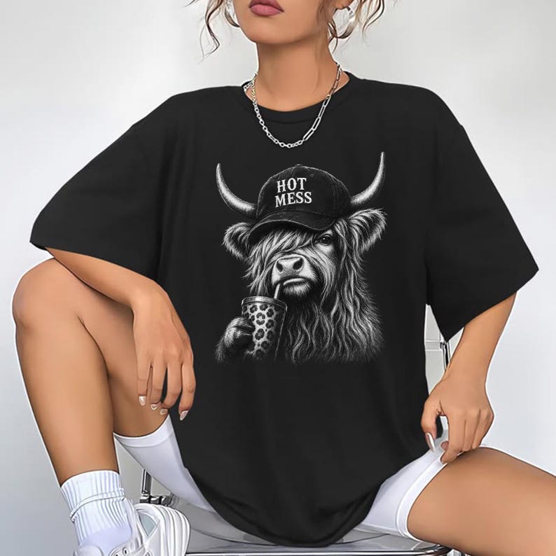 May include: A black short-sleeved t-shirt featuring a graphic of a Highland cow wearing a black baseball cap with the text HOT MESS printed on it. The cow is depicted with long shaggy fur and is holding a leopard-print cup with a straw.