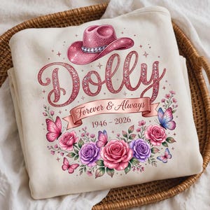 May include: A cream-colored sweatshirt featuring a graphic of a pink glittery cowboy hat above the word Dolly in pink script. Below, a ribbon banner reads Forever & Always 1946-2026, surrounded by pink and purple roses and butterflies.