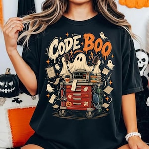 May include: A black t-shirt featuring a graphic of a ghost standing behind a red medical crash cart. The design includes the text "CODE BOO" in cream and orange, along with medical equipment, small ghosts, and a pumpkin icon.