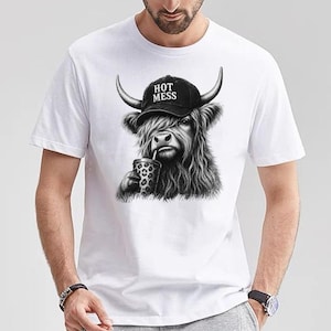 May include: A white short-sleeved t-shirt featuring a black and white graphic of a Highland cow wearing a black baseball cap with the text HOT MESS printed on it. The cow is depicted with long shaggy fur and is holding a leopard print cup.