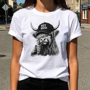 May include: A white t-shirt featuring a black and white graphic of a Highland cow wearing a black baseball cap with the text HOT MESS printed on it. The cow is depicted with long shaggy fur and is holding a leopard print cup with a straw.