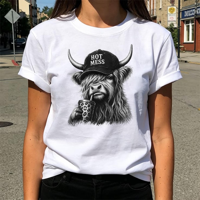 May include: A white t-shirt featuring a black and white graphic of a Highland cow wearing a black baseball cap with the text HOT MESS printed on it. The cow is depicted with long shaggy fur and is holding a leopard print cup with a straw.