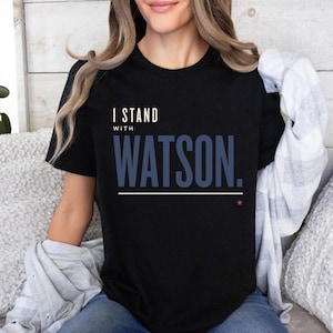 Puede incluir: Una camiseta negra de manga corta con el texto blanco I STAND y el texto azul WATSON. debajo, junto con una fina línea horizontal blanca y una pequeña estrella roja situada bajo el texto en la parte delantera de la prenda.