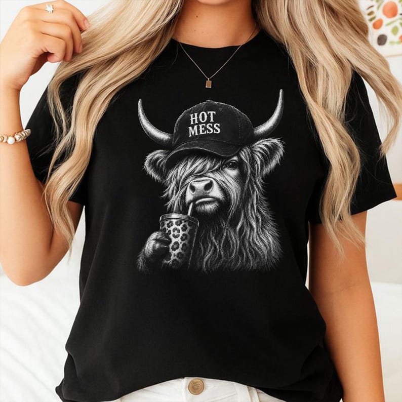 May include: A black t-shirt featuring a graphic illustration of a Highland cow wearing a black baseball cap with the text HOT MESS printed in white. The cow is depicted with long shaggy fur and is holding a leopard print cup with a straw.