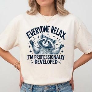 May include: A cream-colored short-sleeve t-shirt featuring a dark blue graphic of a raccoon with its arms raised. The text reads "EVERYONE RELAX I'M PROFESSIONALLY DEVELOPED" in a bold, dark blue font, surrounded by small star accents.