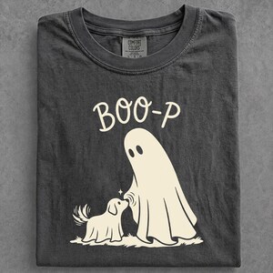 May include: A charcoal gray t-shirt featuring a cream-colored graphic of a ghost and a small dog wearing a ghost costume. The text "BOO-P" is printed in a cream-colored font above the figures. The shirt has a Comfort Colors brand label at the neck.