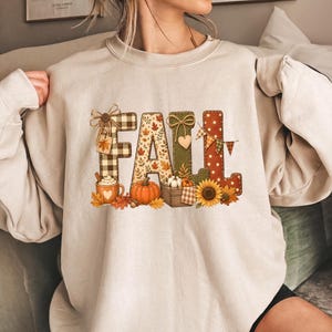Puede incluir: Una sudadera de cuello redondo en color beige con la palabra FALL en letras grandes y decorativas. Las letras están rellenas con patrones de cuadros, hojas de otoño, tela verde y lunares, decoradas con una calabaza, un girasol y una taza.