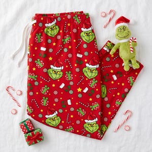 Può includere: Pantaloni da pigiama rossi con un motivo ripetuto del Grinch con cappello da Babbo Natale, scatole regalo verdi, calze rosse e bianche, bastoncini di zucchero e stelle gialle. Un piccolo peluche del Grinch è posizionato sui pantaloni.