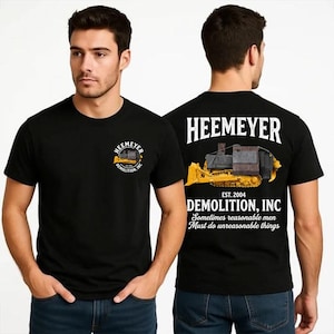 Peut inclure: Un t-shirt noir avec un petit motif sur la poitrine gauche et un grand motif au dos. Les deux graphismes présentent un bulldozer jaune avec le texte HEEMEYER DEMOLITION, INC. EST. 2004 et la phrase Sometimes reasonable men must do unreasonable things.