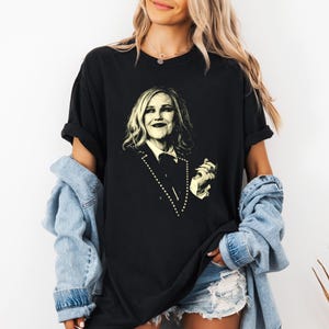May include: Black t-shirt featuring a sepia-toned portrait of a woman. The graphic is centered on the shirt and depicts a person in a suit. The shirt has short sleeves and a relaxed fit. The model is wearing a denim jacket and shorts.