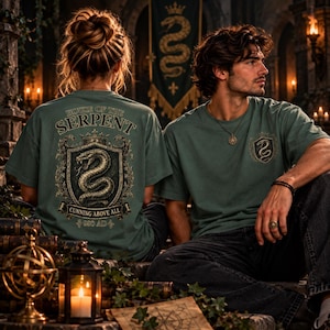 May include: A sage green t-shirt featuring a large graphic on the back with a coiled snake inside a shield, the text "HOUSE OF THE SERPENT" and "CUNNING ABOVE ALL 990 AD". A smaller matching snake shield graphic is printed on the front left chest.