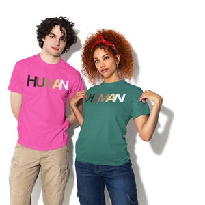 May include: Two people wearing t-shirts with the word "HUMAN" printed across the chest. The word is in a gradient of colors. One shirt is bright pink, the other is teal. The person on the left is wearing khaki pants, and the person on the right is wearing blue jeans.