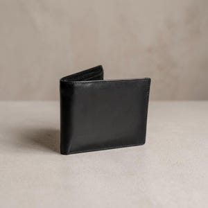 May include: A black leather wallet, open to display the interior. The wallet is rectangular with rounded corners and a smooth, matte finish. The background is a neutral, light color.