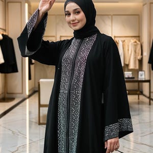 Peut inclure: Abaya noire avec des détails ornés. L'abaya présente une ligne verticale de paillettes scintillantes à l'avant et sur les manches. La femme porte un hijab noir.