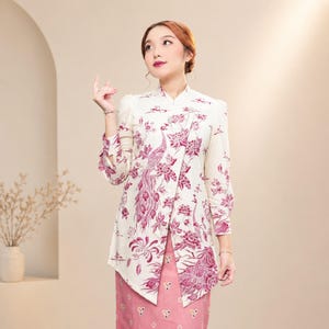 Könnte beinhalten: Ein cremefarbenes Blusen- und rosa Rock-Ensemble. Die Bluse hat einen leuchtenden magenta Pfau- und Blumendruck. Das langärmlige Oberteil hat einen Mandarin-Kragen und ein einzigartiges asymmetrisches Design. Der Rock ist einfarbig rosa.