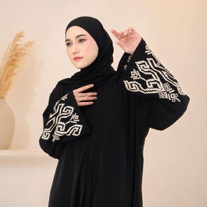 May include: A black abaya with white embroidered patterns on the sleeves. The abaya is worn with a black hijab. The sleeves have a wide, flared design. The garment is made of a soft, flowing fabric.