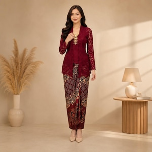 May include: A deep burgundy lace kebaya top with long sleeves and a matching batik sarong skirt featuring intricate geometric and floral patterns in shades of brown, black, and gold. A gold-toned brooch is pinned to the centre of the lace top.