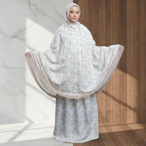 May include: A white prayer garment with a floral pattern and a matching skirt. The garment has a lace trim and a hood. The person in the image is wearing a hijab.