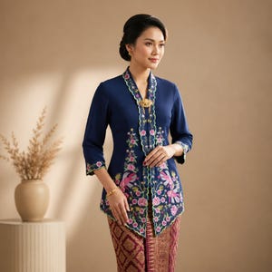 Peut inclure: Un haut Kebaya bleu marine orné de broderies florales roses et vertes, associé à une jupe à motifs. Le haut est orné d'un broche dorée et de bordures délicates. Les manches sont aux trois-quarts. Vêtement traditionnel élégant.