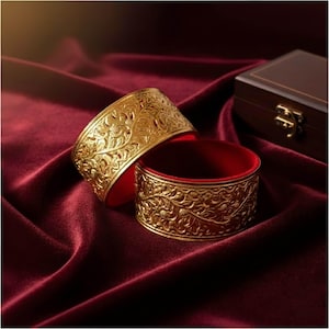 Puede incluir: Dos brazaletes dorados con intrincados diseños florales, sobre una superficie de terciopelo rojo oscuro. Los brazaletes tienen un interior rojo. Una caja de madera con un cierre dorado está en el fondo.