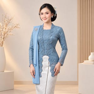 May include: A dusty blue long-sleeved lace kebaya top featuring intricate beadwork and a scalloped hem, paired with a light grey patterned sarong skirt. A matching blue fabric sash is draped over the shoulder, secured with a decorative silver brooch.
