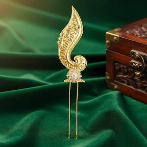 May include: A gold-coloured hair pin with an ornate, leaf-like design. The pin features a decorative, swirling pattern and a central, sparkling jewel. Two long, slender prongs extend from the base.