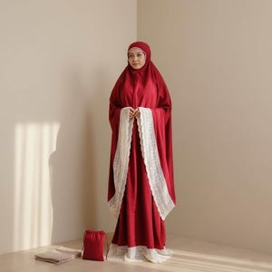 May include: A burgundy prayer garment with white lace edging. The garment includes a matching head covering and a small drawstring bag. A folded beige prayer mat rests on the floor. The woman is standing in a neutral-toned room.