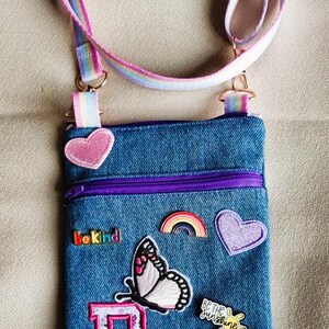 Puede incluir: Bolso cruzado de mezclilla con correa y cremallera arcoíris. La bolsa está decorada con parches que incluyen una mariposa, corazones, un arcoíris y los textos "Be Kind" y "Be the Sunshine".
