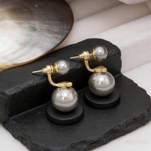 May include: A pair of gold-toned earrings featuring a large, gray pearl at the base and a smaller pearl at the top. The earrings have a curved design and are displayed on a black surface, with a shell in the background.