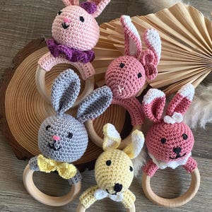 May include: A collection of six handmade crochet bunny rattles in various pastel colors. Each bunny has a wooden ring base and features a unique bow or collar. The bunnies are pink, yellow, gray, and light pink.