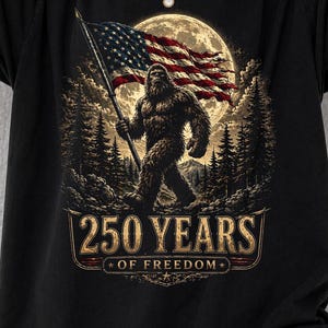 May include: Black t-shirt featuring a Bigfoot graphic holding an American flag in front of a full moon. The design includes the text "250 YEARS OF FREEDOM" in a decorative banner.