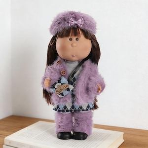 Puede incluir: Muñeca con abrigo, sombrero y pantalón de piel sintética lavanda. La muñeca tiene el pelo largo castaño y lleva un pequeño bolso con la palabra "Smile". La muñeca está sobre un libro.