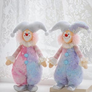 May include: Two plush clown dolls with pastel pink, blue, and purple color-blocked outfits. Each doll has a white collar, a red nose, and a gray jester hat. The dolls have silver shoes and are standing on a white surface.