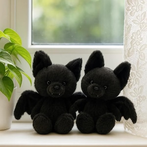 May include: Two plush black bat toys with large ears and small wings sit side-by-side. The toys have small fangs and are positioned on a white surface near a window with a green plant and lace curtain.