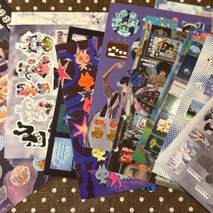 May include: A collection of colorful stickers featuring various designs, including anime characters, cartoon figures, and abstract art. The stickers are arranged in a stack, showcasing a variety of shapes and sizes, with a brown polka dot background.