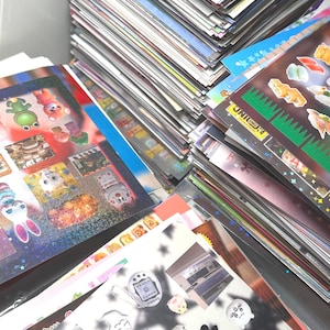 May include: A large stack of assorted sticker sheets featuring various colorful designs, including cartoon animals, food items, digital pet devices, and anime-style illustrations. The sheets have diverse finishes, ranging from holographic to matte.