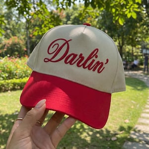 May include: A beige and red baseball cap with the word "Darlin'" embroidered in red script across the front. The cap has a red brim and a beige crown, perfect for casual wear.