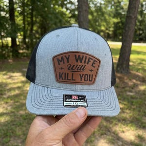 May include: A gray and black trucker hat with a brown leather patch that reads "MY WIFE WILL KILL YOU." The hat has a curved brim and a mesh back. The brand is Richardson.