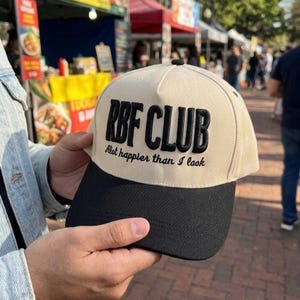 May include: A beige and black baseball cap with the text "RBF CLUB" in bold black letters. Below the text is the phrase "A lot happier than I look" in a smaller font. The cap has a curved brim.