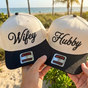 May include: Two baseball caps, one cream and black, with the words "Wifey" and "Hubby" in black script. Each cap has a black brim and a small tag. The caps are a mid-profile fit.