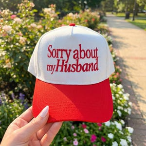 May include: A white and red baseball cap with the embroidered text "Sorry about my Husband" in red. The cap has a red brim and a white crown, with a red button on top. The hat is a fun accessory.